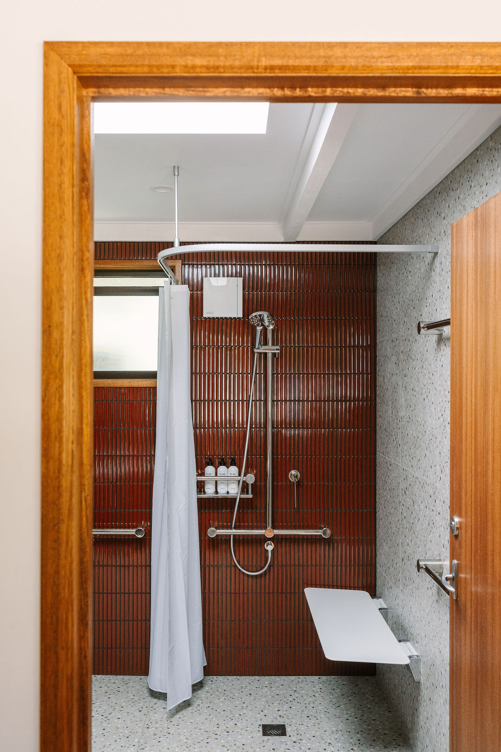 Accessible bathroom with tiled walls and speckled flooring, featuring a walk-in shower with a white bench seat, handheld shower head and grab bars, and a privacy curtain; Accommodation Bright, Mid Century Stays, Victoria Alpine