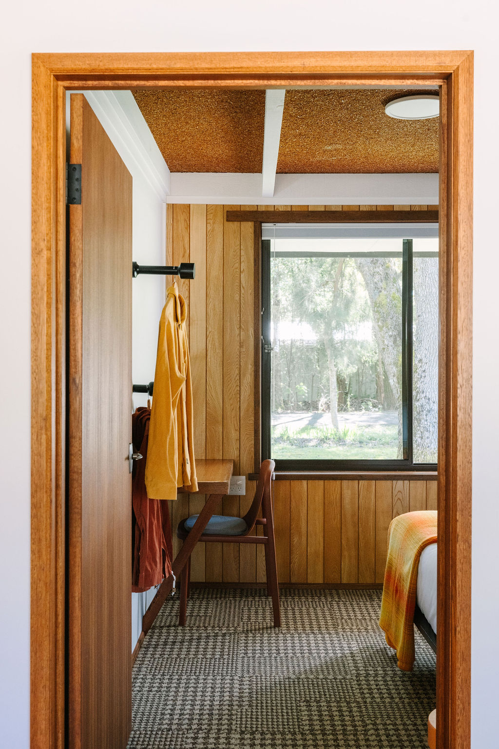 Interior view of an accessible bedroom at Bomen Harrietville, featuring wood-paneled walls, a carpeted floor, a cosy bed with a warm orange blanket, a desk and chair by a large window looking out to trees, and clothing hooks; Accommodation Bright, Mid Century Stays, Victoria Alpine