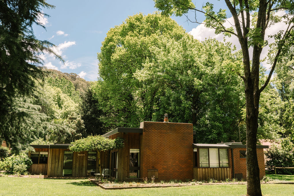 Exterior view of Blackwood Lodge at Bomen Harrietville showing a mid-century brick cabin with large windows among shady trees and lush lawn; Accommodation Bright, Mid Century Stays, Victoria Alpine