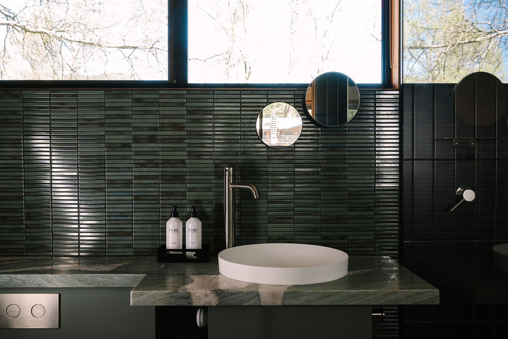 Bathroom vanity at Bomen Harrietville featuring a green tiled backsplash, stone countertop with round vessel sink, brass faucet, and round mirror; Accommodation Bright, Mid Century Stays, Victoria Alpine