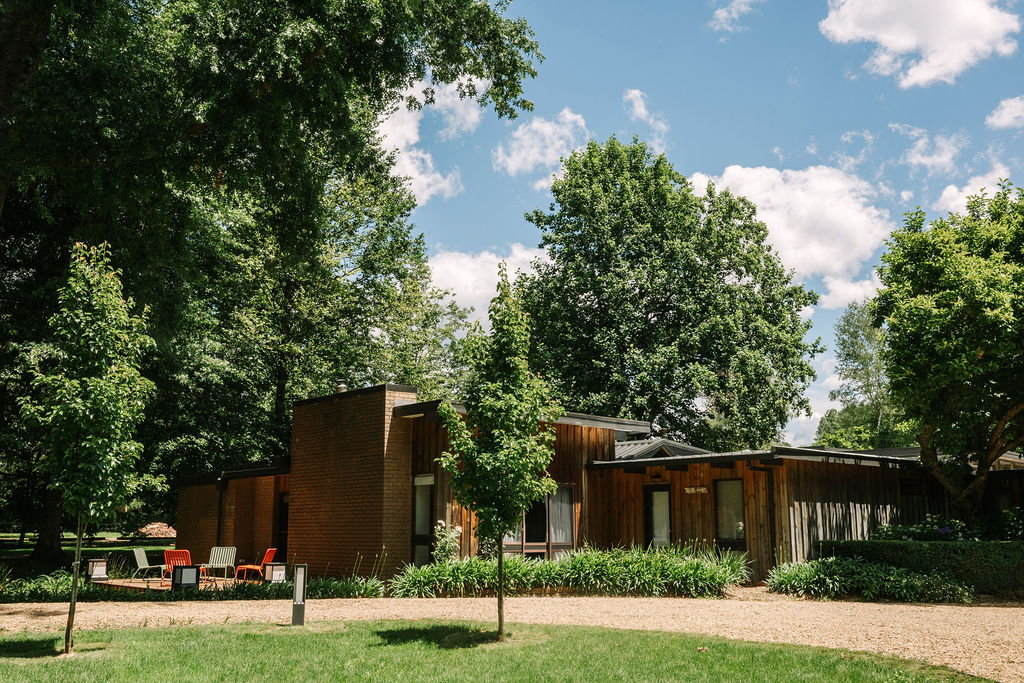 Exterior view of Myrtle Lodge at Bomen Harrietville, a mid-century style cabin with timber facade set among lush trees and gardens, with a gravel driveway and lawn area; Accommodation Bright, Mid Century Stays, Victoria Alpine