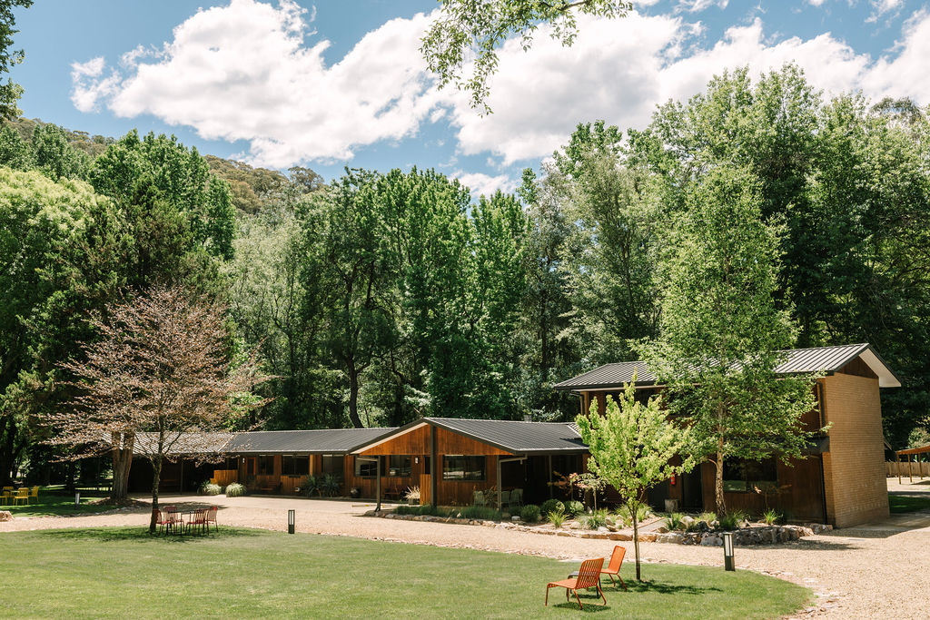 Exterior view of the 2-bedroom cabin at Bomen Harrietville (N2) showing modern timber buildings with pitched roofs and covered verandas, nestled among tall trees and grassy lawns with gravel pathways; Accommodation Bright, Mid Century Stays, Victoria Alpine