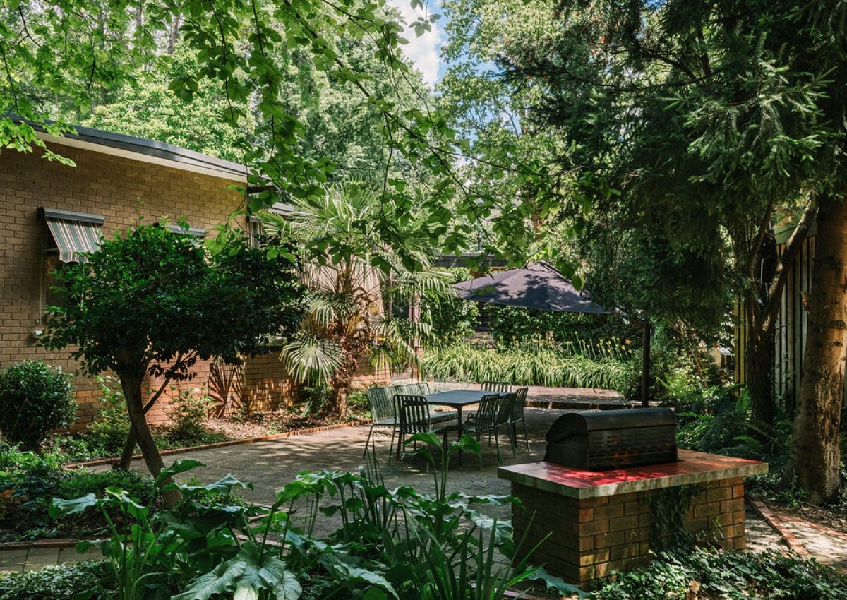Lush courtyard at Blackwood Lodge featuring a paved patio area with outdoor dining table and chairs, a built-in barbecue grill, and verdant gardens under tall trees; Accommodation Bright, Mid Century Stays, Victoria Alpine